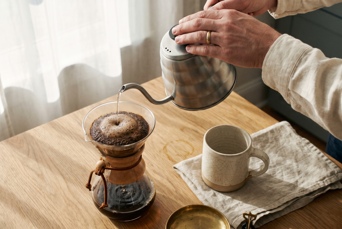 Manos vertiendo agua sobre una Chemex con el café en bloom, junto a un pocillo de cerámica