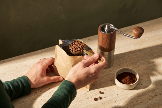 Manos sacando café en grano de una bolsa kraft junto a un molino manual y café molido